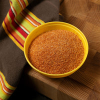 A yellow bowl filled with The Original French Fry Seasoning—a fine, orange-brown blend—rests on a wooden surface beside a brown cloth with red, yellow, and orange stripes.