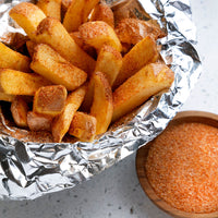 Thick-cut potato fries coated with French Fry Seasoning - The Original sit in a foil-lined bowl, with a small wooden bowl beside them filled with extra French Fry Seasoning - The Original.