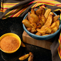 A blue bowl of golden, seasoned potato wedges sits on a wooden board next to a yellow bowl of French Fry Seasoning - The Original, with striped napkins in the background.