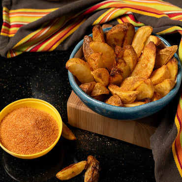 A blue bowl of golden, seasoned potato wedges sits on a wooden board next to a yellow bowl of French Fry Seasoning - The Original, with striped napkins in the background.