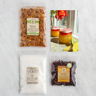 Three clear spice bags labeled Mango Tango, Mango, and Hibiscus Rosé are arranged around a recipe card displaying two tropical drinks with a lime wedge and red flowers on a sunny windowsill—the perfect setting for crafting a mango hibiscus cocktail with the vibrant Tropical Bay Breeze (Kit).
