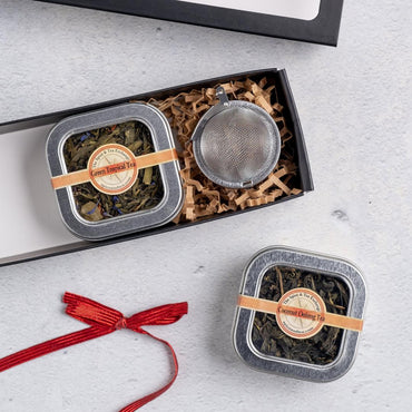The Tropical Tea Tin Gift Box includes Green Tropical Tea, Coconut Oolong Tea, a metal infuser, and crinkle paper in a gift box. A red ribbon rests nearby on the light gray surface—ideal for tropical tea enthusiasts.
