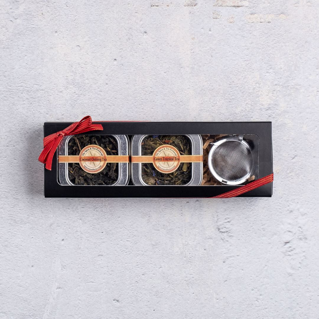 The Tropical Tea Tin Gift Box features two tins of tropical teas, including coconut oolong, and a metal infuser in a black rectangular box with a red ribbon, displayed on a light grey textured surface.