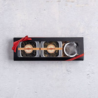 The Tropical Tea Tin Gift Box features two tins of tropical teas, including coconut oolong, and a metal infuser in a black rectangular box with a red ribbon, displayed on a light grey textured surface.