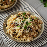 A close-up of a plate of risotto with mushrooms, parmesan cheese, and fresh parsley garnish, served on a striped napkin.