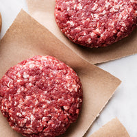 Two raw ground meat burger patties with visible fat marbling, placed on torn brown parchment paper over a light surface.