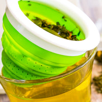 Close-up of a green Tuffy Steeper Infuser with holes, filled with loose tea leaves, resting on a glass mug of yellow brewed tea. Ideal travel tea infuser for enjoying fresh flavors anywhere; available in blue, green, purple, and fuchsia.