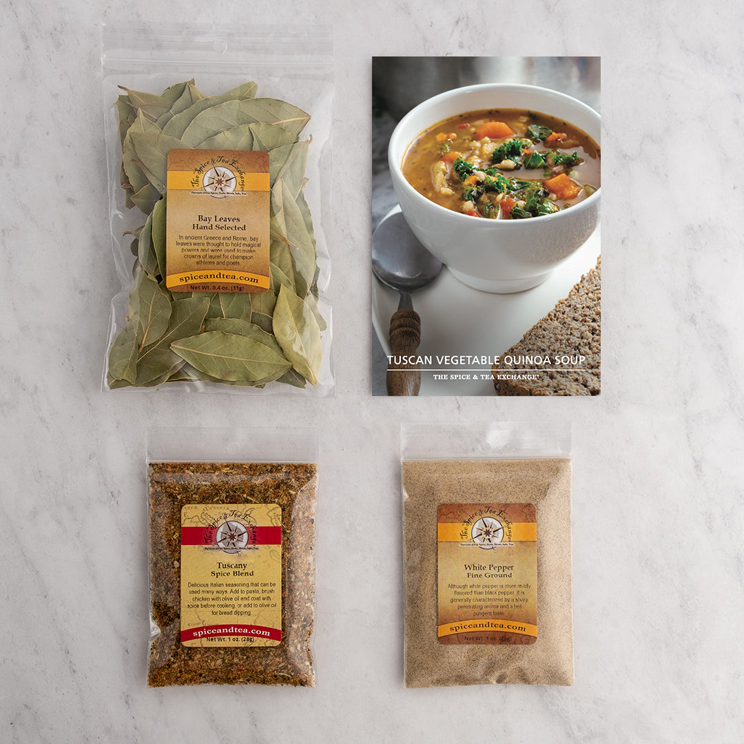 Three clear spice packets (bay leaves, Tuscany Spice Blend, white pepper) sit on a marble surface next to a recipe card featuring a bowl of Tuscan Vegetable Quinoa Soup (Kit) with a spoon.