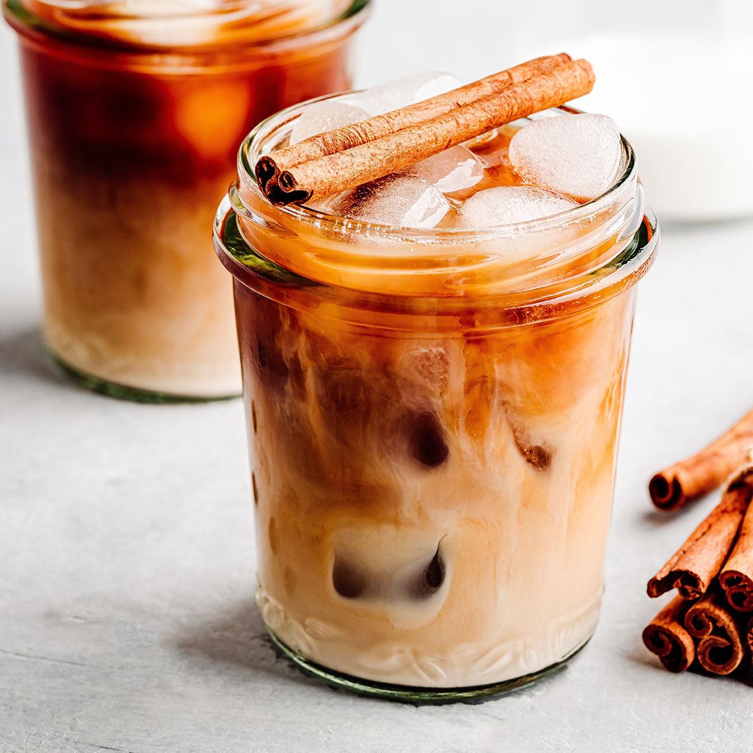 A glass of Victorian Chai Tea swirls with cream and floral notes, topped with two cinnamon sticks and ice cubes. Another glass of iced Victorian Chai Tea and loose cinnamon sticks rest in the background.