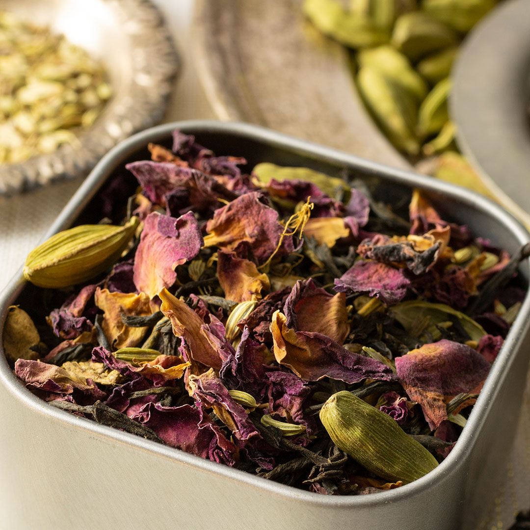 A close-up of Victorian Chai Tea shows a metal container filled with dried rose petals, green cardamom pods, and tea leaves. In the background, extra cardamom pods and seeds on blurred trays add delicate floral notes to the scene.