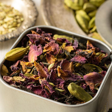 A close-up of Victorian Chai Tea shows a metal container filled with dried rose petals, green cardamom pods, and tea leaves. In the background, extra cardamom pods and seeds on blurred trays add delicate floral notes to the scene.
