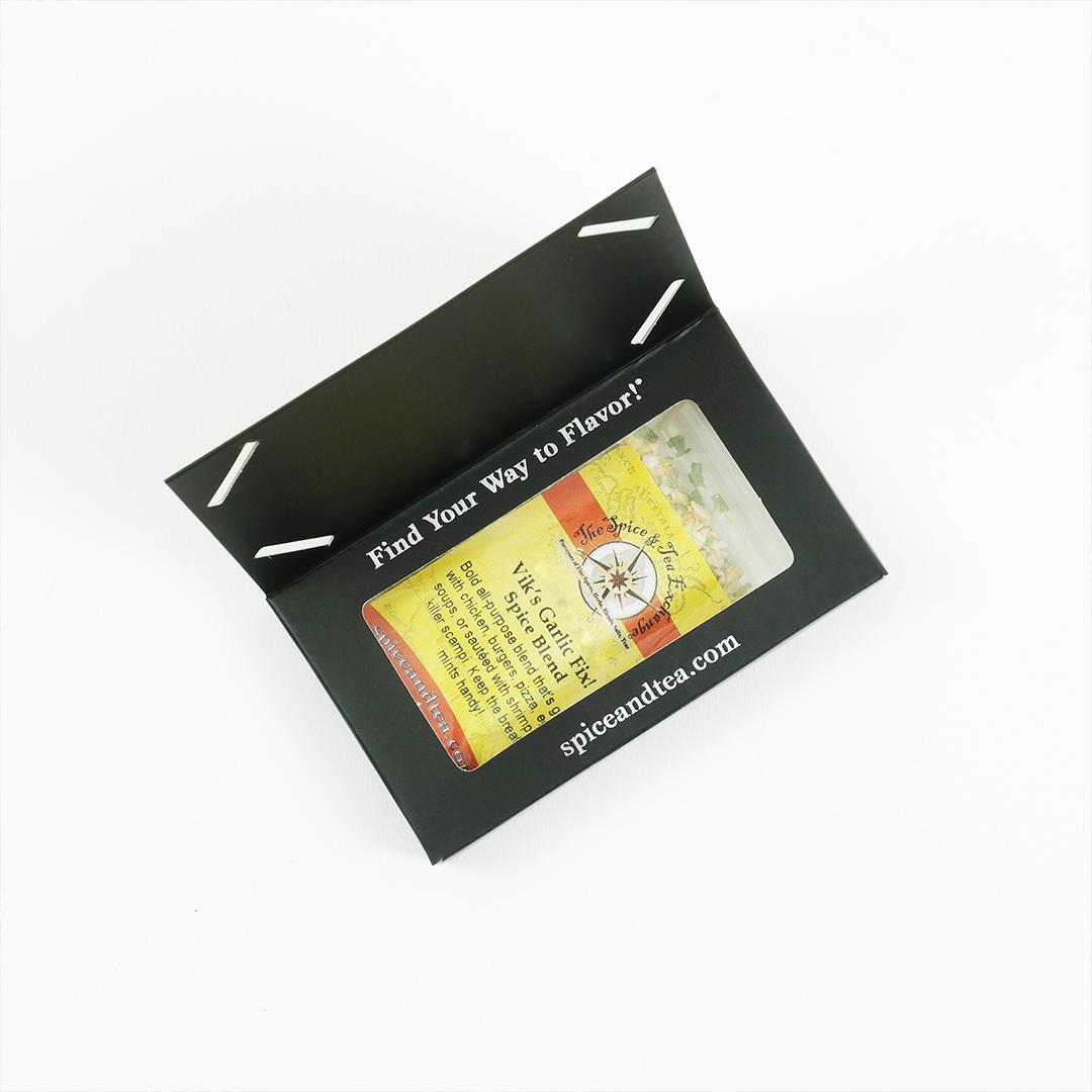 A sleek black card envelope with white text, “Find Your Way to Flavor!” and “spiceandtea.mom,” holds a Vik's Garlic Fix Spice Blend - Barter Box from The Spice & Tea Exchange—visible through a clear window—perfect as a gift sampler.