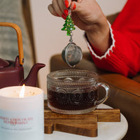 Person holding a tea infuser over a glass mug with a lit candle and teapot in the background.