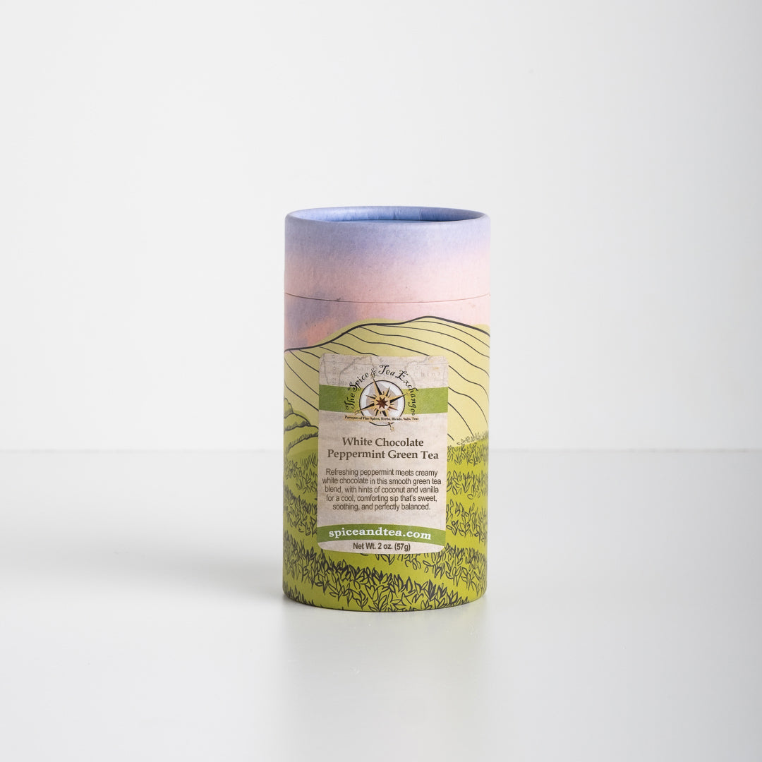 A cylindrical tea canister featuring a purple and green landscape design, labeled White Chocolate Peppermint Green Tea from spiceandtea.mom, rests on a plain white surface. Subtle notes of green tea and coconut vanilla are present.