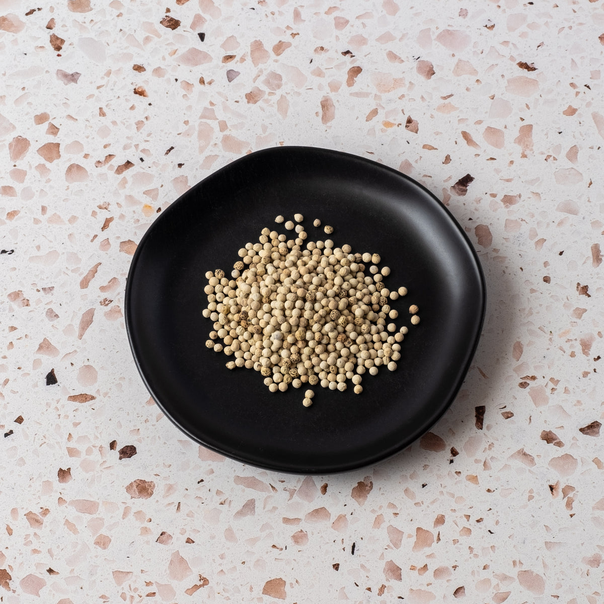 A small pile of White Peppercorn, a prized spice for cream sauces, sits on a round black plate placed on a light speckled terrazzo surface.