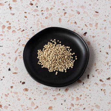 A small pile of White Peppercorn, a prized spice for cream sauces, sits on a round black plate placed on a light speckled terrazzo surface.