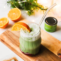 A glass of Cranberry Orange Matcha Latte, featuring ceremonial matcha and an orange slice, rests on a wooden board with a halved orange, a green tin from the kit, and festive decor in the background.