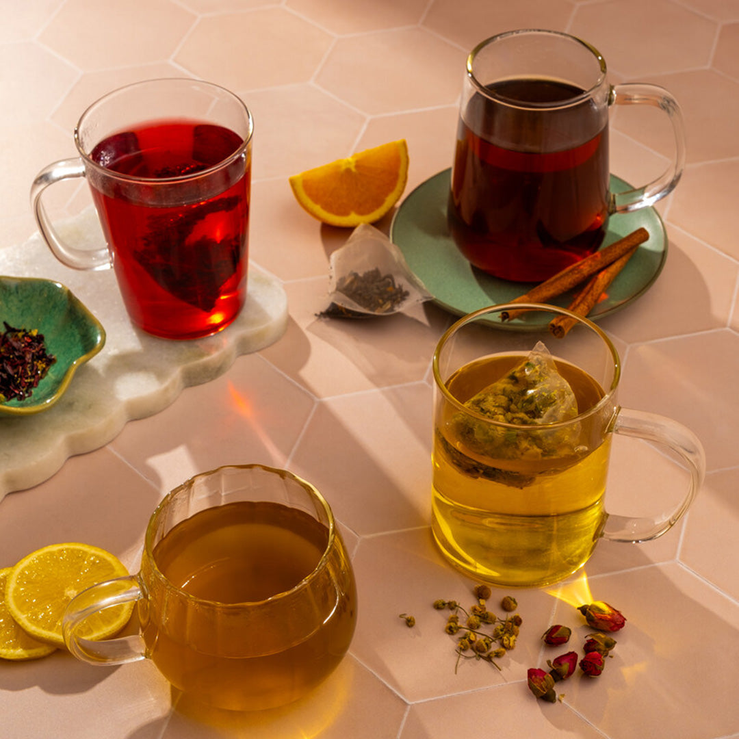 Three glass mugs with organic teas—red, dark brown, and yellow—from the 7 Day Organic Tea Cleanse sit on a light tiled surface, surrounded by lemon slices, orange wedge, cinnamon sticks, dried flowers, and tea bags for a perfect cleanse.