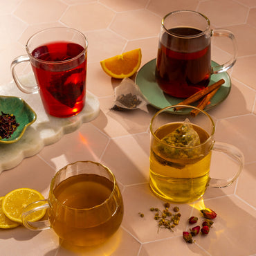 Three glass mugs with organic teas—red, dark brown, and yellow—from the 7 Day Organic Tea Cleanse sit on a light tiled surface, surrounded by lemon slices, orange wedge, cinnamon sticks, dried flowers, and tea bags for a perfect cleanse.