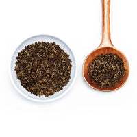 A white bowl and a wooden spoon, both filled with ground black pepper, sit side by side on a white background, ready to be paired with Porcini Mushroom Sea Salt for enhanced flavor.