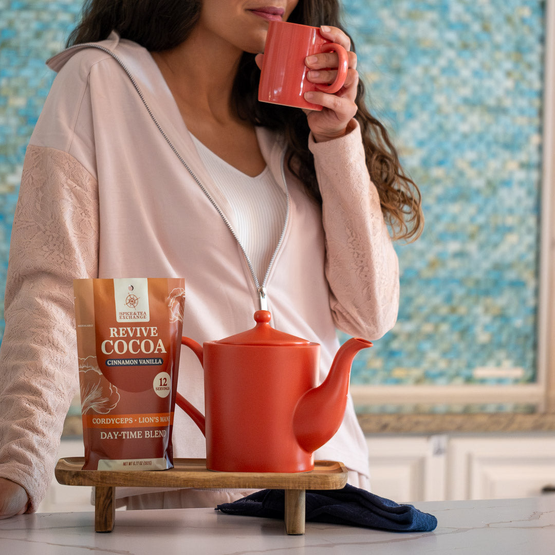 A woman in a light pink hoodie holds a red tea mug infuser near her mouth. In front of her is a Cactus Flower Teapot, a packet labeled Revive Cocoa, and a small wooden tray on the kitchen counter with a blue towel underneath.